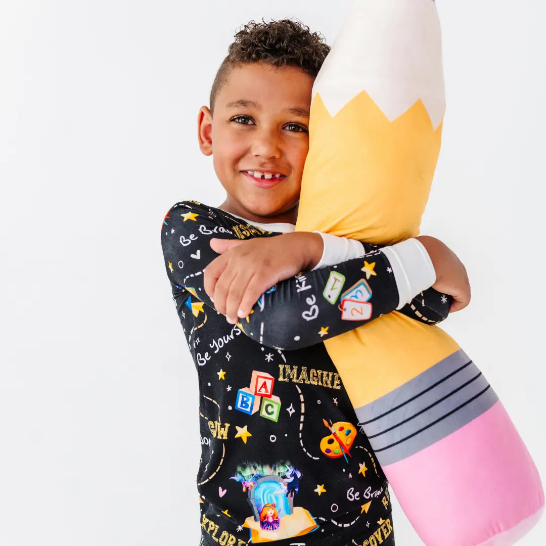 A child in black school-print pajamas smiling while hugging a large plush pencil pillow.